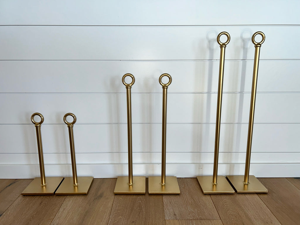  gold-finished metal stand holders with circular top rings and square bases, arranged in ascending size order on a wood floor against a white shiplap wall.