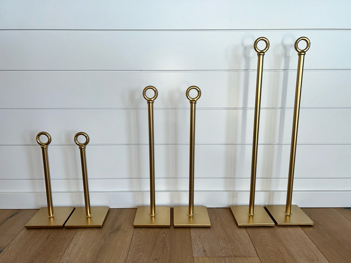  gold-finished metal stand holders with circular top rings and square bases, arranged in ascending size order on a wood floor against a white shiplap wall.
