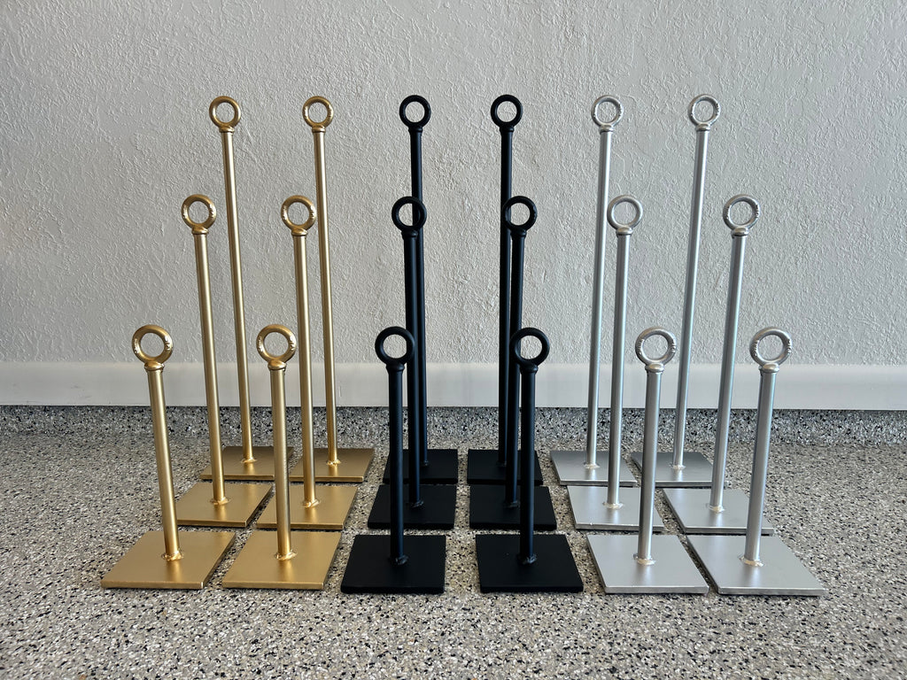 Three sets of metal display stands in gold, black, and silver, arranged side-by-side on a speckled floor against a textured wall.